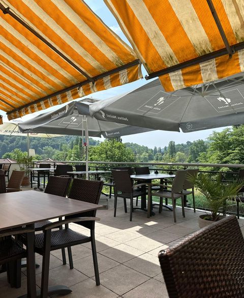 Ein gemütlicher Außenbereich eines Cafés mit Tischen und Stühlen. Die Terrassenmarkisen in Orange und Weiß bieten Schutz vor der Sonne.