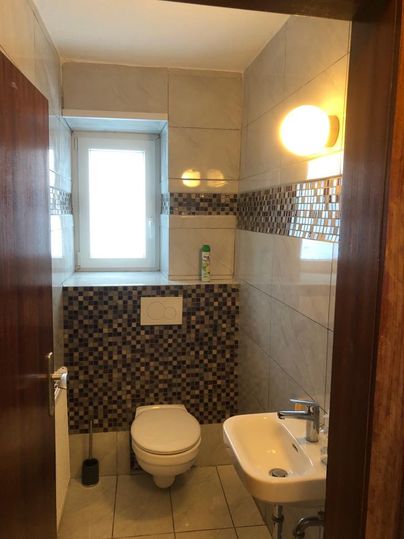 Ein modernes Badezimmer mit einem weißen WC und einem kleinen Waschbecken. An den Wänden sind helle Fliesen und ein Fenster sorgt für natürliches Licht.