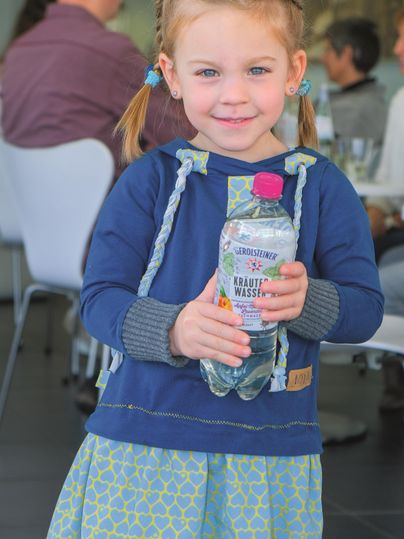 A girl with braids is holding a bottle of Gerolsteiner herbal water. In the background, people are sitting in a visitor center.