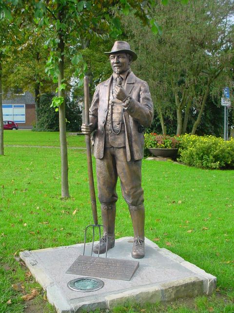 A bronze statue of a man with a hat holding a pitchfork. The statue stands in a green park with trees and flowers.