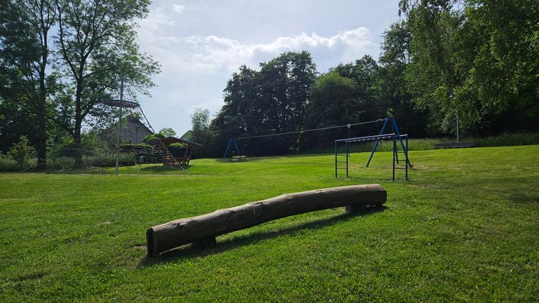 Ein schöner Spielplatz mit grüner Wiese und Schaukeln. Im Hintergrund sind Bäume und ein Spielgerät zu sehen.