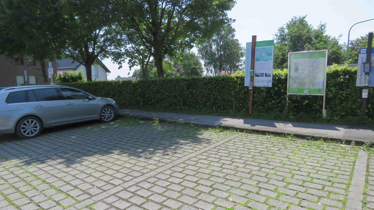 Een parkeerplaats met een geparkeerde zilveren auto. Op de achtergrond zijn informatieschilderen en een groene haag te zien.