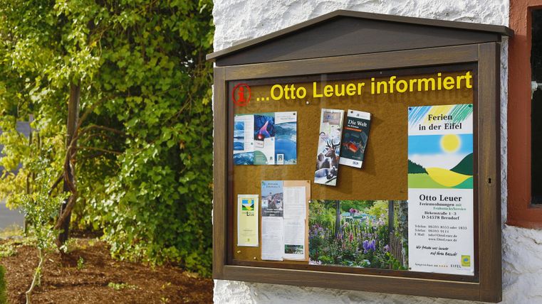 Ein Info-Brett an einer Wand mit verschiedenen Flyern und Karten. Es steht "Otto Leuer informiert" in gelben Buchstaben.