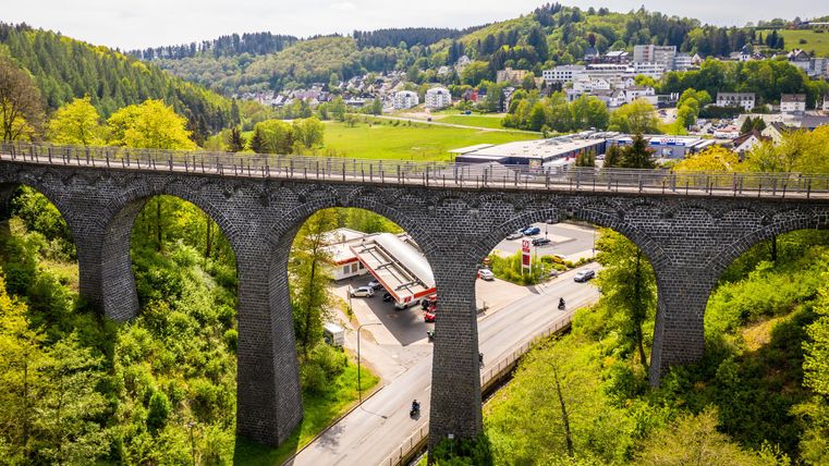 Eine beeindruckende Brücke über ein Tal mit grünen Bäumen. Darunter verläuft eine Straße, und im Hintergrund sieht man ein kleines Dorf.
