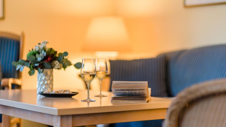 Ein gemütlicher Wohnbereich mit einem Tisch, auf dem zwei Weingläser und eine kleine Blumenvase stehen. Im Hintergrund ist eine Couch und eine Lampe zu sehen.