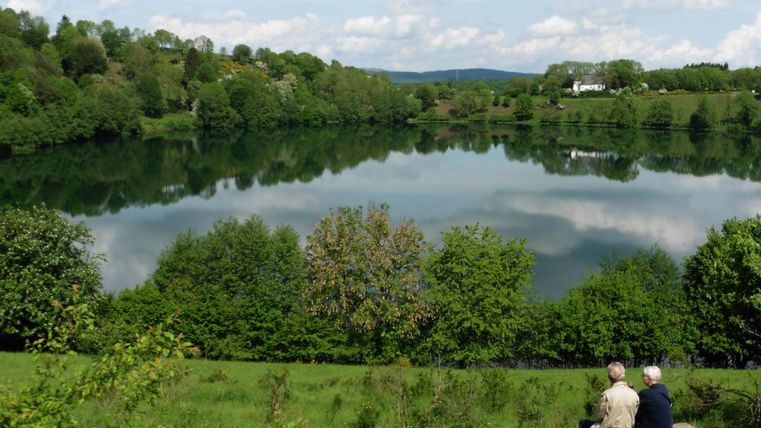 Ein ruhiger See, umgeben von grünen Bäumen und sanften Hügeln. Zwei Personen sitzen auf einer Bank und genießen die Aussicht.