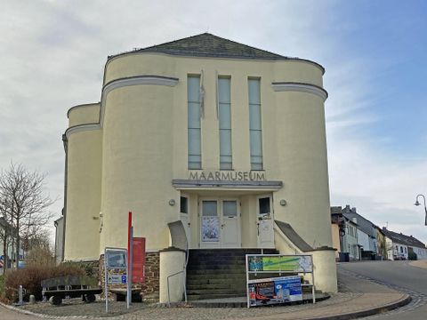 Das Maarmuseum ist ein modern gestaltetes Gebäude mit einer auffälligen, runden Fassade. Vor dem Eingang stehen Informationen und Plakate zur Ausstellung.