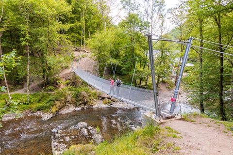 A suspension bridge over a small river, surrounded by dense green forests. Two people are crossing the bridge and enjoying nature.