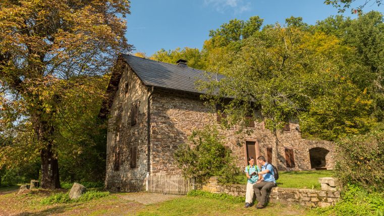 Zwei Personen sitzen vor einem alten Steinhaus im Grünen.