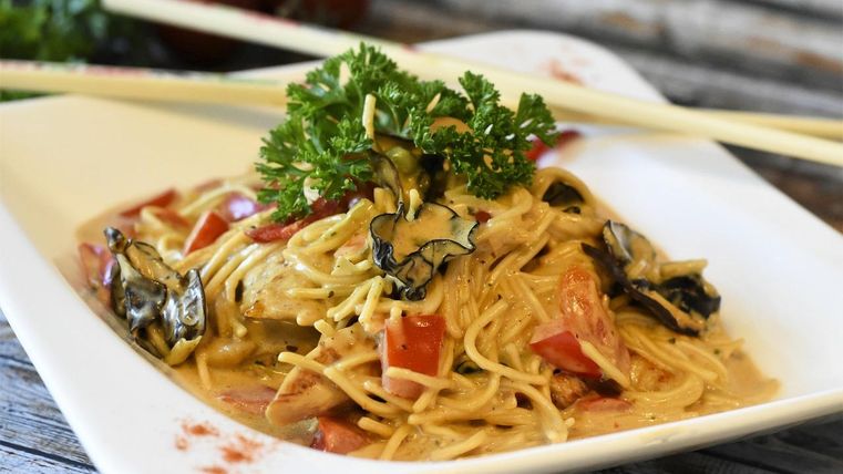 A plate with a high edge, on which an Asian noodle dish is presented with various vegetables and herbs. Chopsticks are resting on the edge of the plate.
