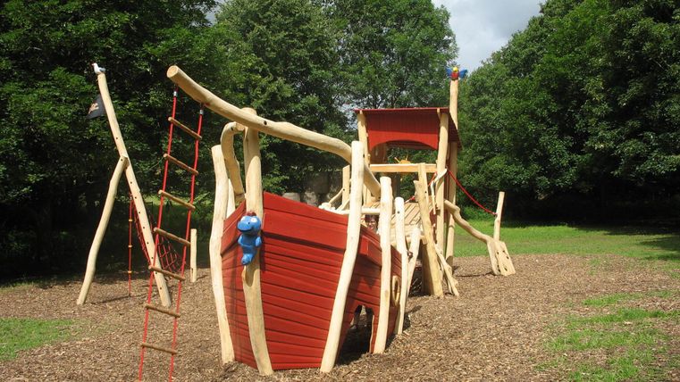 Ein Spielplatz mit einem Holzschiff mit Rutsche und Klettergerüst. Umgeben von grünen Bäumen und einer naturbelassenen Fläche.