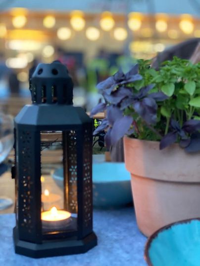 Een stijlvolle lantaarn staat op een tafel, omringd door verse kruiden in een bloempot. Op de achtergrond zijn zachte lichten te zien die een ontspannen sfeer creëren.