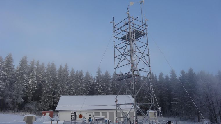 Ein hoher Turm steht neben einem kleinen Gebäude in einer winterlichen Landschaft. Der Boden ist mit Schnee bedeckt und Bäume sind im Hintergrund sichtbar.