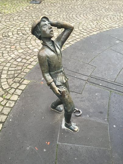 A bronze statue of a boy standing with one hand on his head and looking up. The ground is made of stone slabs and gravel.