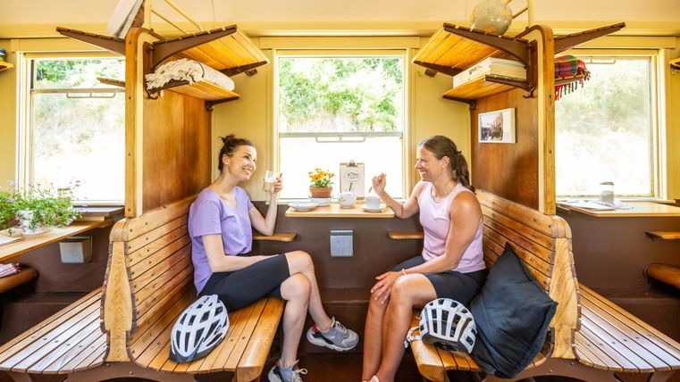 Twee vrouwen zitten in een lichte kamer op een houten bank. Ze genieten van drankjes en lachen met elkaar.