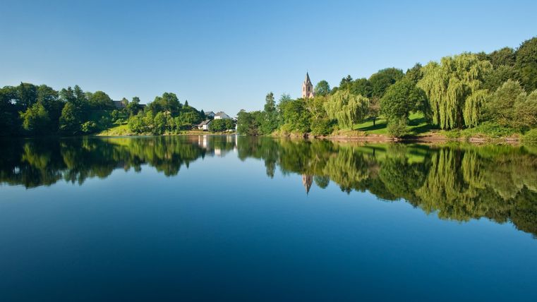 Ein ruhiger See mit spiegelndem Wasser und grünen Ufern. Der Himmel ist klar und blau.
