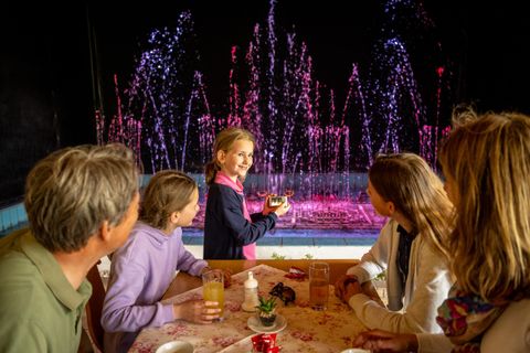 A family is sitting at a table and looking at an illuminated water fountain organ. A child is pointing at something on a smartphone.