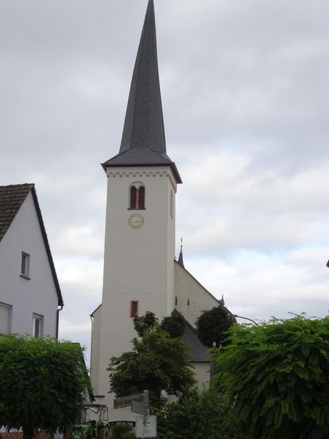A church building with a tall, pointed tower. Surrounded by low houses and green plants.