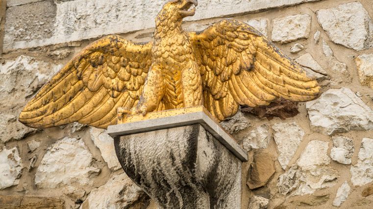 A golden eagle with outstretched wings sits on a stylish pillar. The statue is mounted on a stone wall.