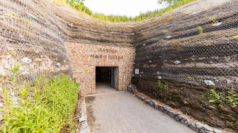 Ein Eingang zu einem Stollen, umgeben von Erdwänden und Pflanzen. Über dem Zugang steht der Schriftzug „Magma Stollen“.