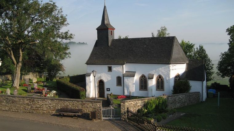In the middle of the picture, you can see the small whitewashed chapel. The roof is covered with black slate. On the left side, there is a church tower with a pointed roof, also covered in black.
At the top of the church tower, you can see a weather vane. The chapel is surrounded by a churchyard that is framed by a rubble stone wall.