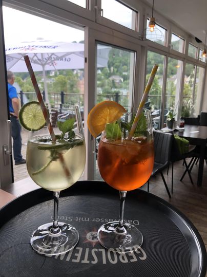Two cocktails with lime and orange slices on a tray in a bistro, in the background a terrace with a sunshade.