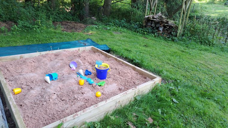 A sandpit in a garden, filled with sand and toys. Surrounded by green grass and trees in the background.