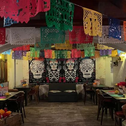 Ein festlich geschmückter Raum mit bunten Papierflaggen und einem Wandbild von Totenschädeln. Die Tische sind hübsch gedeckt und tragen zur lebendigen Atmosphäre bei.