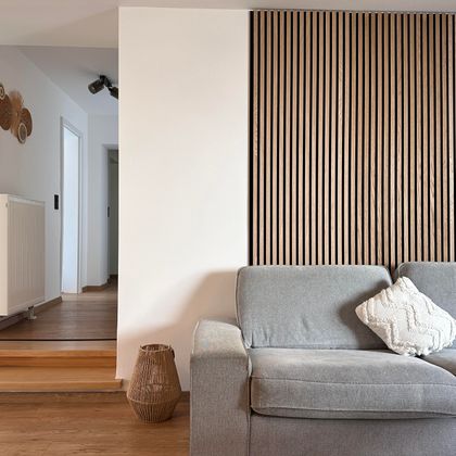 Ein modernes Wohnzimmer mit einem grauen Sofa und einer dekorativen Holzvertäfelung an der Wand. Der Boden ist aus hellem Holz und schafft eine einladende Atmosphäre.