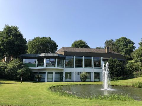 Ein modernes Gebäude mit großen Fenstern liegt auf einer grünen Wiese. Im Vordergrund befindet sich ein Teich mit einem Springbrunnen.