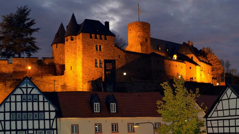 Eine beeindruckende Burg beleuchtet in der Dämmerung. Im Vordergrund sieht man charmante Fachwerkhäuser.