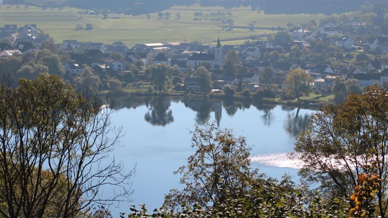 A tranquil lake surrounded by green landscape and small villages. The mirrored water surface reflects the beauty of nature.