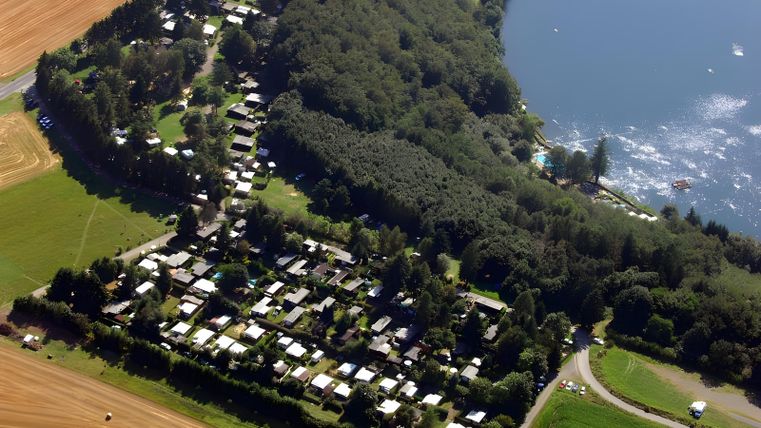 Een luchtfoto van een vakantiepark aan de oever van een meer, omringd door bomen en velden. De indeling van de campers en erven is duidelijk herkenbaar.