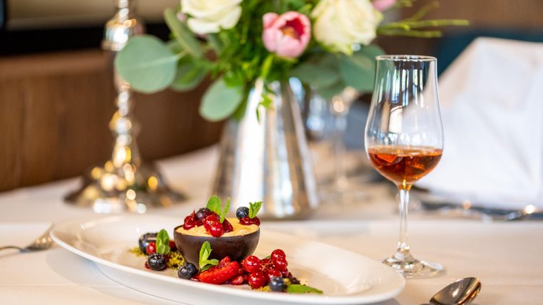 Ein elegantes Gericht mit Schokoladendessert und frischen Beeren. Im Hintergrund steht ein Glas Wein und eine Blumenvase.