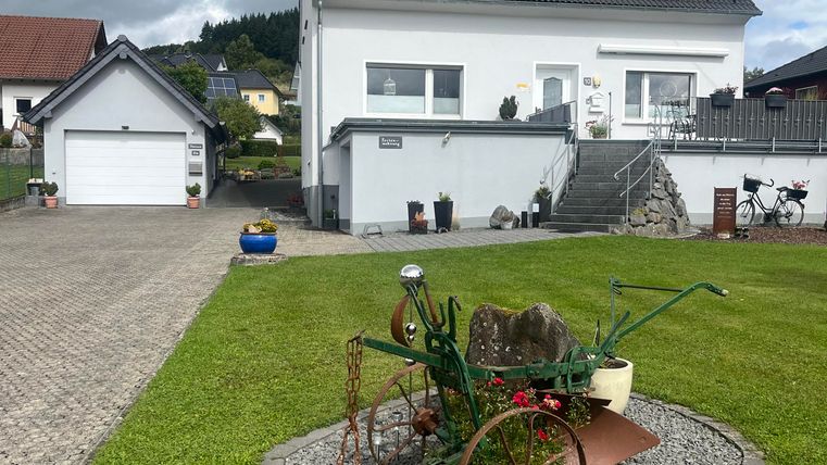 Ein modernes Haus mit Solarpanelen auf dem Dach steht in einer grünen Umgebung. Im Vordergrund befindet sich ein dekorativer Garten mit einem alten Wagen.