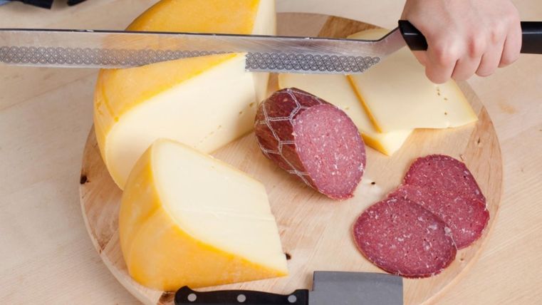 Ein rundes Holzbrett, auf dem einhalbes Laib Käse liegt und mit einem großen Messer in Scheiben geschnitten wird. Daneben ein weiteres Käsemesser und eine Salami.
