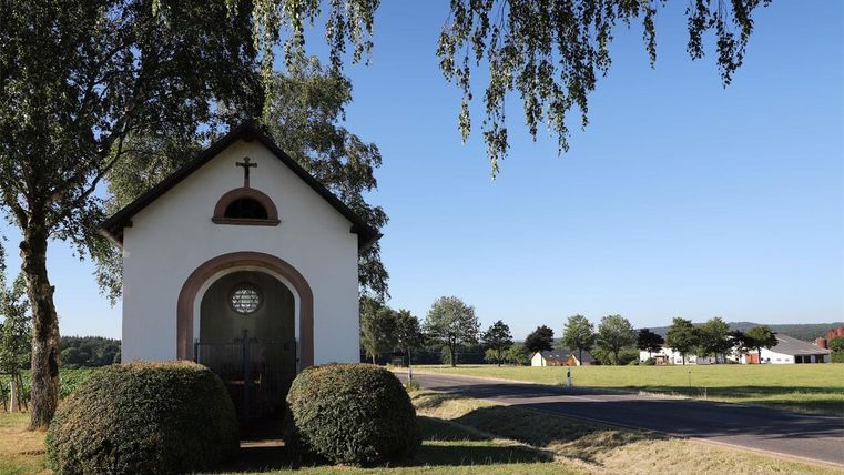 A small chapel with a cross above the door stands beside a road. Surrounded by trees and meadows, it offers a tranquil atmosphere.