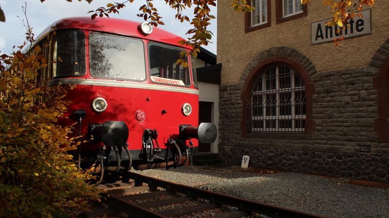 A red train is standing on the tracks in front of a train station building. The surroundings are surrounded by autumn leaves in warm colors.