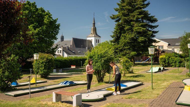 Een midgetgolfbaan met meerdere banen en groenvoorzieningen. Op de achtergrond is een kerk en blauwe lucht te zien.