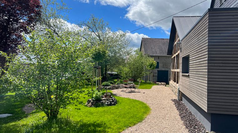 Ein schöner Garten mit viel Grün und blühenden Bäumen. Im Hintergrund ist ein modernes Gebäude zu sehen, umgeben von einem Kiesweg.