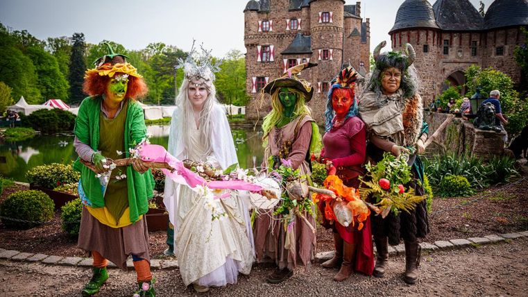 Eine Gruppe von fantasievoll verkleideten Menschen steht vor einem Schloss. Sie tragen bunte Kostüme und halten dekorierte Stäbe, umgeben von einer malerischen Landschaft.