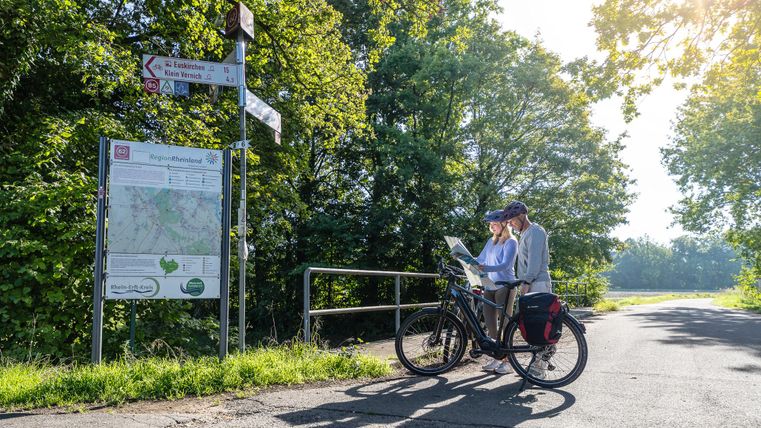 Twee fietsers staan bij een wegwijzer in het groen en bestuderen een kaart. De zonnige dag en de dichte bomen creëren een ontspannen sfeer.