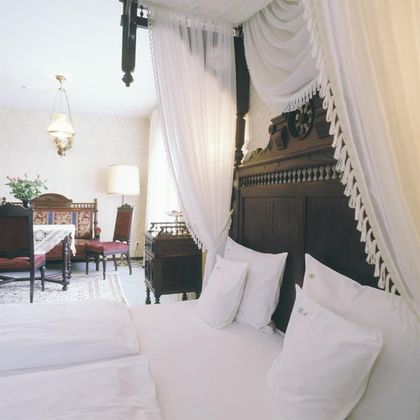 A cozy bedroom with a large, decorative bed and elegant bedding. In the background, there is a stylishly furnished living area with a table and chairs.