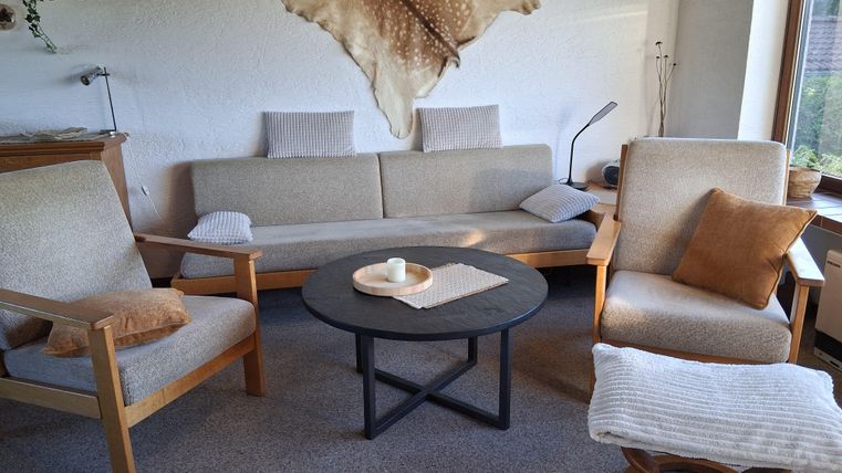Ein gemütliches Wohnzimmer mit einer beigen Couch und zwei Stühlen. Ein runder Tisch steht in der Mitte, und eine dekorative Wandgestaltung ist sichtbar.