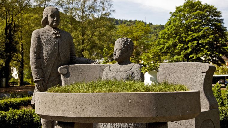 Eine Steinfigur zeigt einen Mann und eine Frau, die an einem runden Tisch sitzen. Um sie herum ist eine grüne Landschaft mit Bäumen.