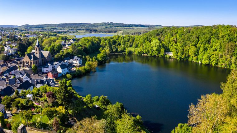 Ein malerischer Teich umgeben von grünen Wäldern und kleinen Häusern. Im Hintergrund steht eine Kirche, die die Landschaft wunderschön ergänzt.