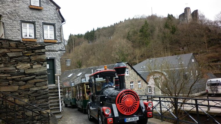 An old town with stone houses and a small tourist train. In the background, hills and a part of a castle ruin are visible.