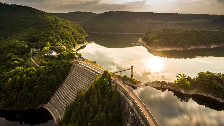 Eine beeindruckende Staudammlandschaft mit einem ruhigen See und sanften Hügeln. Das Licht der untergehenden Sonne spiegelt sich im Wasser wider.