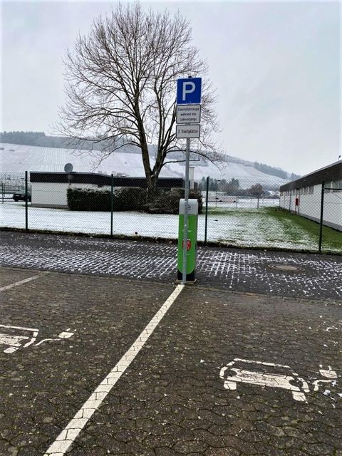 Ein Parkplatz mit einem Parkschild steht vor einer verschneiten Landschaft. Im Hintergrund sind Bäume und ein Zaun zu sehen.