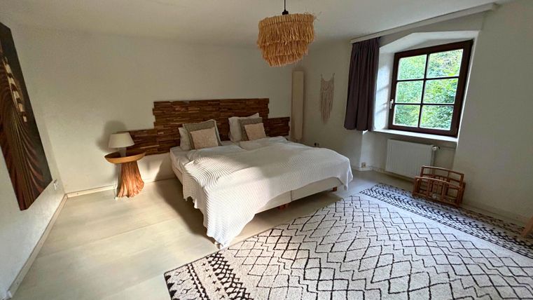 A modern bedroom with a large bed and decorative pillows. The room features natural materials and a window with a view of the outdoors.
